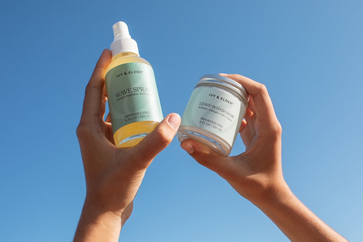 Two hands holding Ivy & Elder hair care products against a clear blue sky: a Wave Spray on the left and a jar of Leave-in hair Mask on the right.