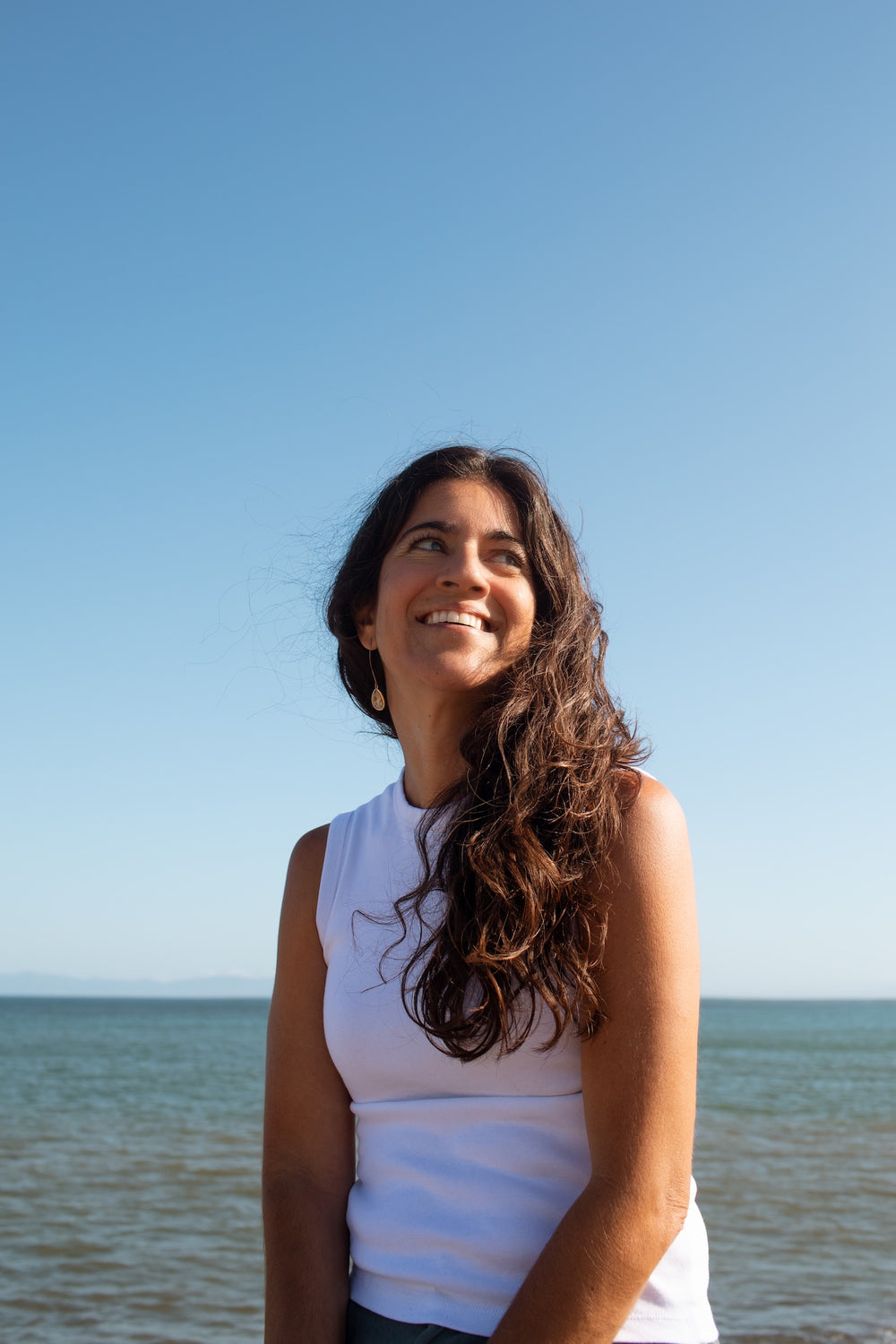 A smiling woman with long dark wavy hair stands outdoors by the water under a clear blue sky, wearing a white sleeveless top.