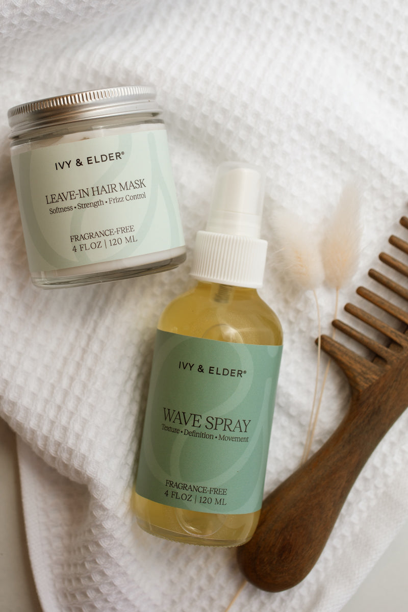 Two Ivy & Elder hair care products arranged on a white textured towel with a wooden hair comb: a Leave-In Hair Mask jar and a Wave Spray bottle. The products are styled with natural props.