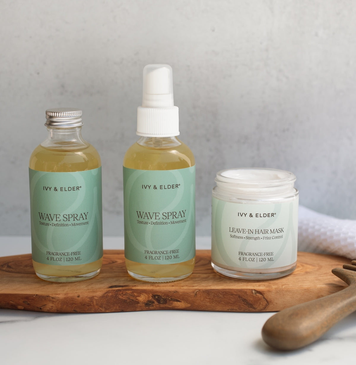 Three hair care products from Ivy & Elder are arranged on a wooden board against a light gray background: a bottle of Wave Spray refill, a regular bottle of Wave Spray and and a jar of Leave-in hair mask next to a comb.