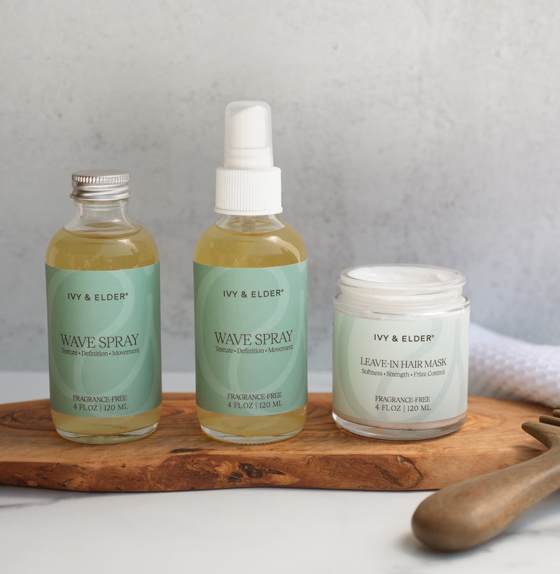 Three hair care products from Ivy & Elder are arranged on a wooden board against a light gray background: a bottle of Wave Spray refill, a regular bottle of Wave Spray and and a jar of Leave-in hair mask next to a comb.
