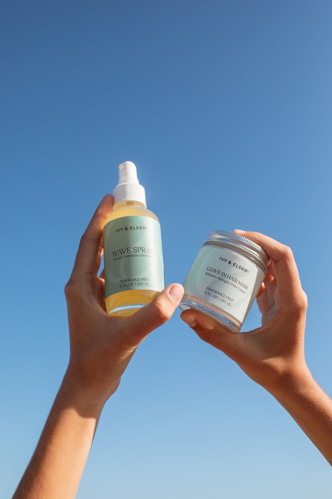 Two hands holding Ivy & Elder haircare products against a clear blue sky: a light green spray bottle  and a matching jar with a white lid.