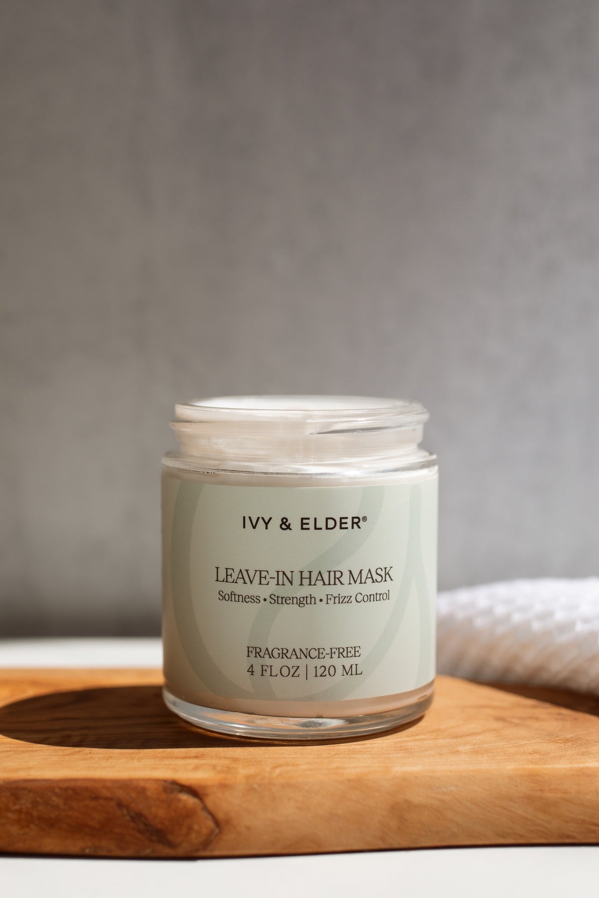 A jar of Ivy & Elder Leave-In Hair Mask sits on a wooden board against a neutral gray background.