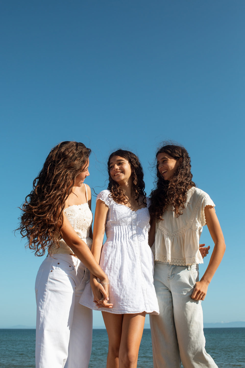 Three young women standing outdoors by the ocean under a clear blue sky, each wearing a white outfit. 