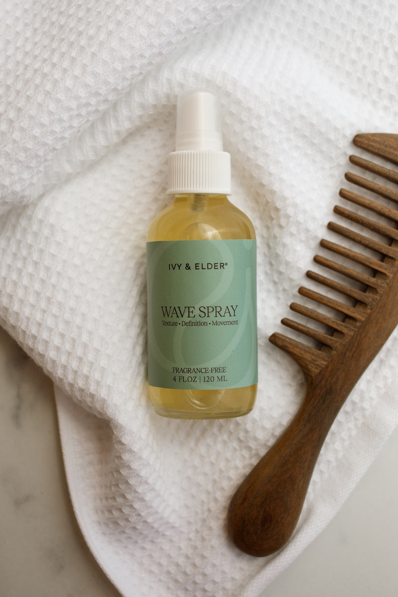 A bottle of Ivy & Elder Wave Spray with a white spray cap and a light green label, placed on a white textured waffle towel next to a wooden comb.