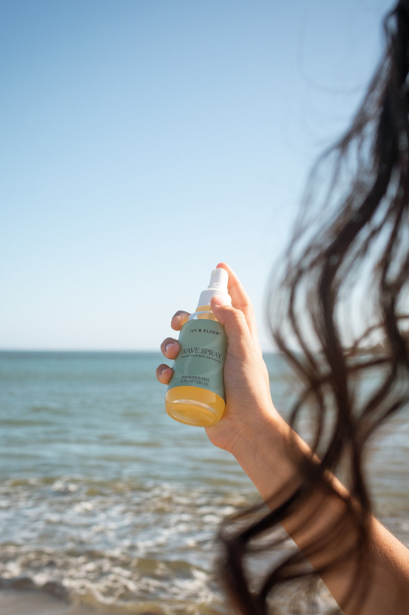 A hand holds a bottle of Wave Spray in front of a beach background, with the person's dark hair visible in the foreground and the ocean and blue sky behind.