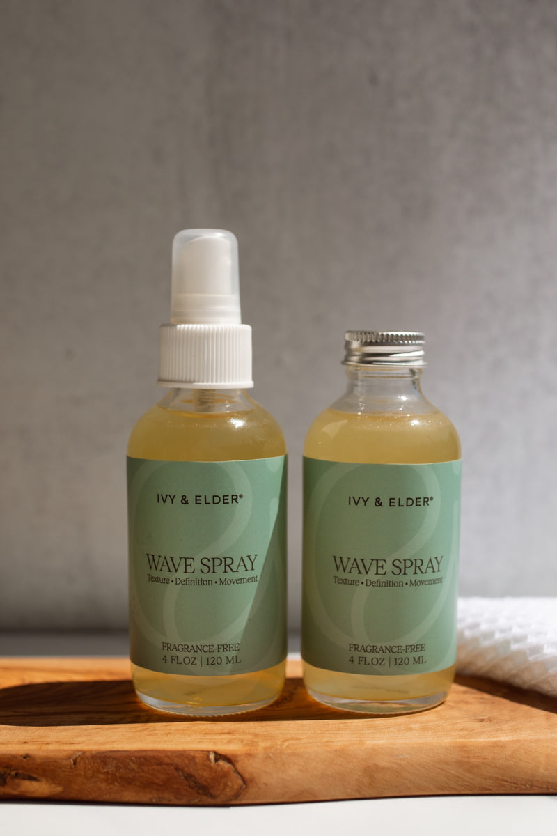 Two bottles of Ivy & Elder Wave Spray, one in a clear pump bottle with a white cap and one in a clear bottle with a silver cap, both containing a pale yellow liquid and featuring light green labels, displayed on a wooden board against a textured gray background.